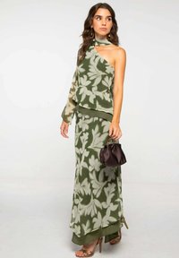 Mujer vestida con un vestido de un solo hombro en verde oliva y beige con estampado floral y una manga larga, sosteniendo un pequeño bolso marrón oscuro y llevando tacones dorados.