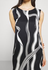 Svart ärmlöst kläde med ett vågigt mönster i vitt, tillverkat av ett mjukt material. Anmärkningsvärt detalj: en liten etikett på sidan.