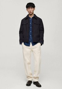 Marinejacke mit Knopfverschluss, darunter ein blauer Denim-Hemd, cremefarbene Hose und schwarze Stiefeletten. Einfaches, strukturiertes Design und kontrastierende Farben.