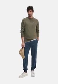 Sudadera verde oliva con cuello redondo, pantalones jogger azules y gorra beige. Zapatillas blancas con detalles en gris y sutil branding en la ropa.