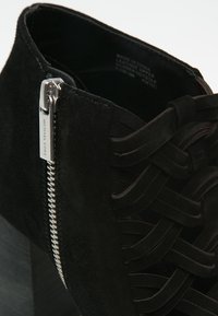 Gros plan d’une bottine noire en daim avec fermeture éclair argentée portant l’étiquette Michael Kors et des brides en daim croisées à l’avant.