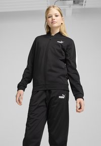 Survêtement noir en tissu lisse, comprenant une veste zippée avec des accents blancs et un logo, accompagnée de pantalons fuselés assortis.