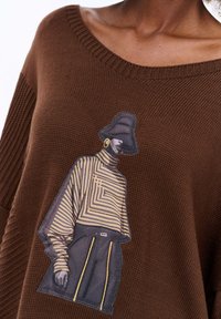 Bruine gebreide trui met een brede halslijn. Bevat een afbeelding van een figuur in gestreepte kleding, met een hoed en oorbel, geplaatst op de voorkant.