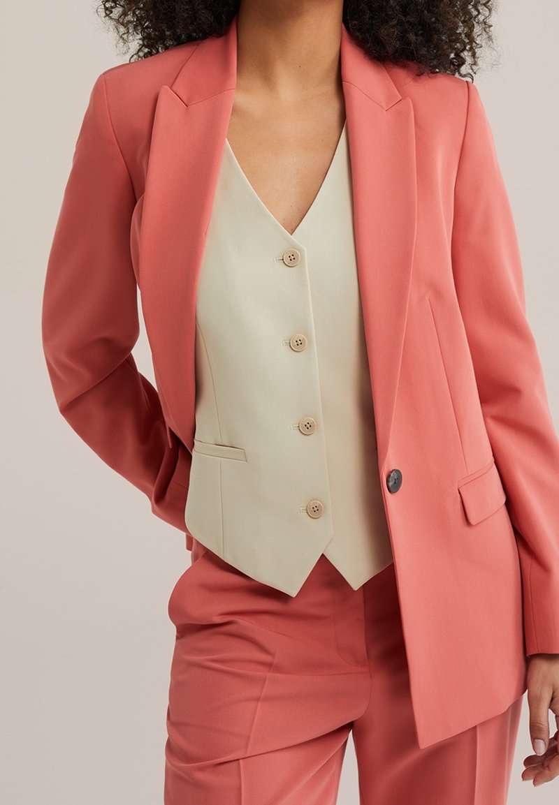 Femme portant un costume rose saumon avec un gilet crème boutonné, debout avec une main derrière le dos.