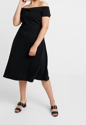 Mujer con vestido midi negro sin hombros, mangas cortas y sandalias negras con tacón de bloque y punta abierta.