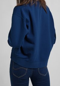 Sudadera azul marino con un corte relajado, dobladillo acanalado y hombros caídos, combinada con jeans de mezclilla oscuros que tienen bolsillos traseros curvados.