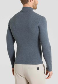 Polo a maniche lunghe in tessuto grigio scuro, con colletto alla coreana e silhouette aderente, indossato con pantaloni beige chiaro, che mostrano un dettaglio di maglia testurizzata.