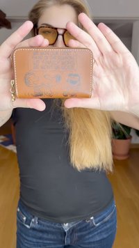 Portafoglio in pelle marrone con grafica cartoon in rilievo che raffigura personaggi e testo. Chiusura con zip e anello portachiavi inclusi.