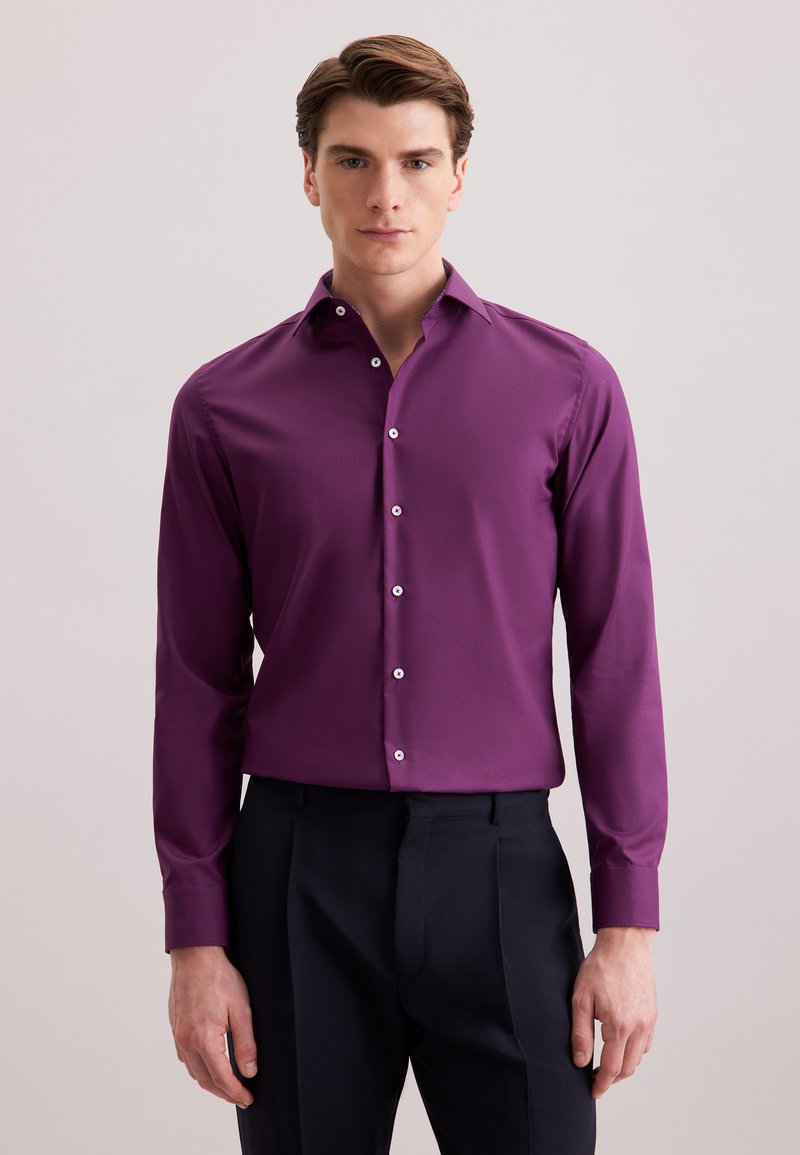 Giovane uomo in piedi, che indossa una camicia aderente viola con bottoni e pantaloni neri, su uno sfondo chiaro e semplice.