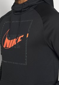 Felpa nera Nike con un grande logo rosso, motivi geometrici e una consistenza morbida. Include un cappuccio con coulisse e maniche a contrasto.