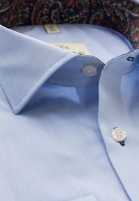 Lichtblauwe shirt met een gladde, gelijkmatige textuur, voorzien van een gemusterde donkere kraagvoering en twee witte knopen bij de nek.