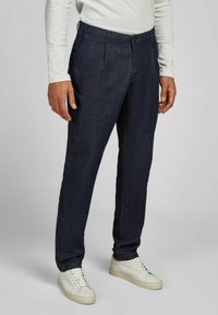 Man in een donkerblauwe rechte broek en witte sneakers, staande tegen een effen lichtgrijze achtergrond.