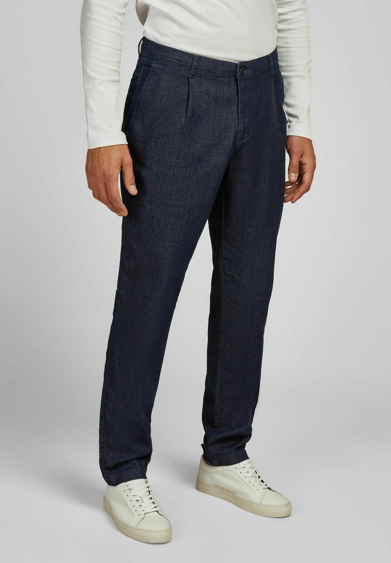 Man in een donkerblauwe rechte broek en witte sneakers, staande tegen een effen lichtgrijze achtergrond.