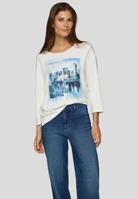 Weißes langärmeliges Oberteil mit einem blauen Grafikdruck einer Stadtsilhouette. Kombiniert mit blauen Jeans mit weitem Bein und schafft so einen lässigen Look.