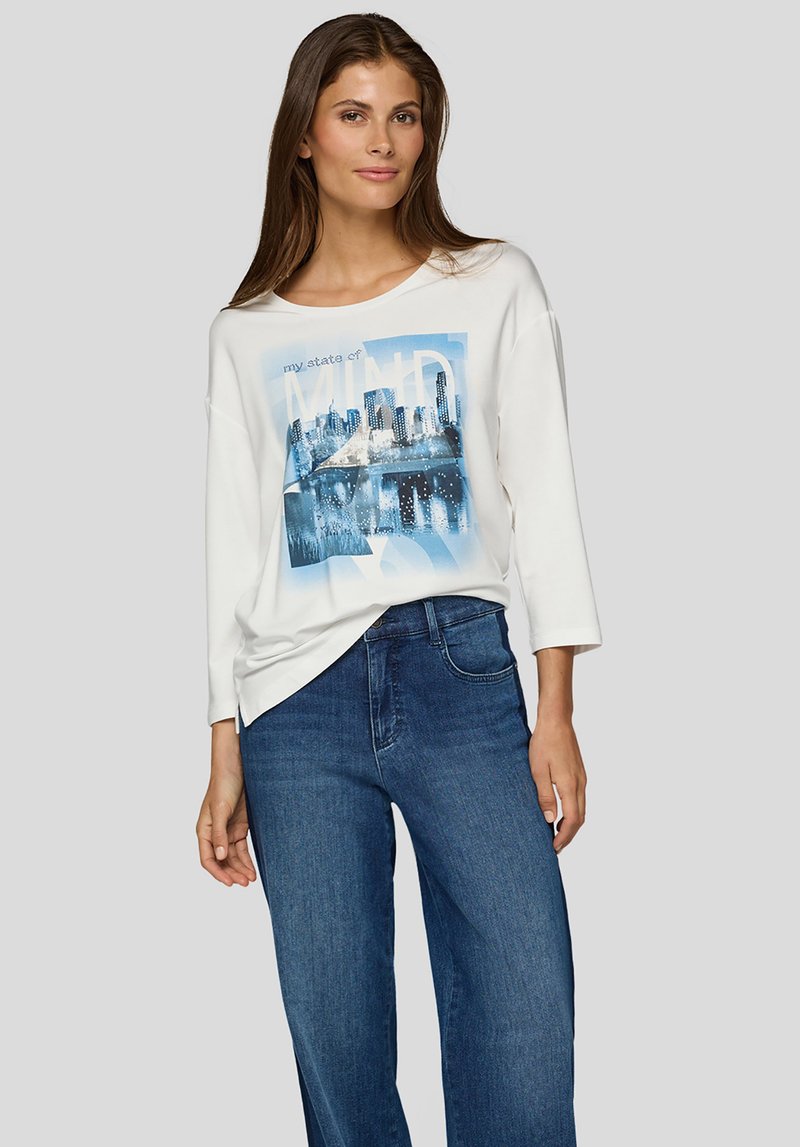 Weißes langärmeliges Oberteil mit einem blauen Grafikdruck einer Stadtsilhouette. Kombiniert mit blauen Jeans mit weitem Bein und schafft so einen lässigen Look.