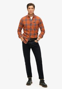 Camisa de franela a cuadros en naranja y azul con mangas largas, combinada con jeans oscuros y botas negras de cordones. Estilo casual con un corte relajado.
