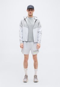 Ανοιχτό γκρι αδιάβροχο με ασημένιο σχέδιο, γκρι μπλουζάκι και αθλητικό σορτς με το λογότυπο "NIKE"; συνδυασμένα με ανοιχτά αθλητικά παπούτσια και κάλτσες.