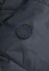 Czarna pikowana kurtka wykonana z tkaniny z subtelnym geometrycznym wzorem oraz okrągłym gumowym logiem marki na części piersiowej, o gładkiej fakturze.