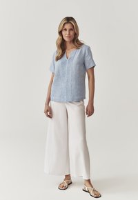 Top di lino azzurro chiaro a maniche corte con scollo a V e pieghe verticali; abbinato a pantaloni bianchi a gamba larga e sandali neri.