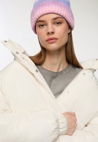 Biała kurtka puchowa o miękkiej teksturze i zapięciu na napy, noszona na szarym podkoszulku. Modelka ma na sobie różowo-niebieską dzianinową czapkę w paski.