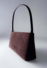 Hnědá semi�šová trapezoidní kabelka s jedním zaobleným koženým uchy na jednoduše šedém pozadí.