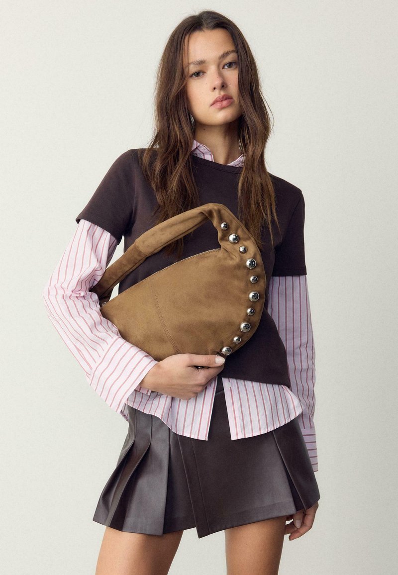 Jeune femme portant une chemise rayée superposée et un haut noir avec une jupe plissée, tenant un sac en daim marron avec des clous argentés.