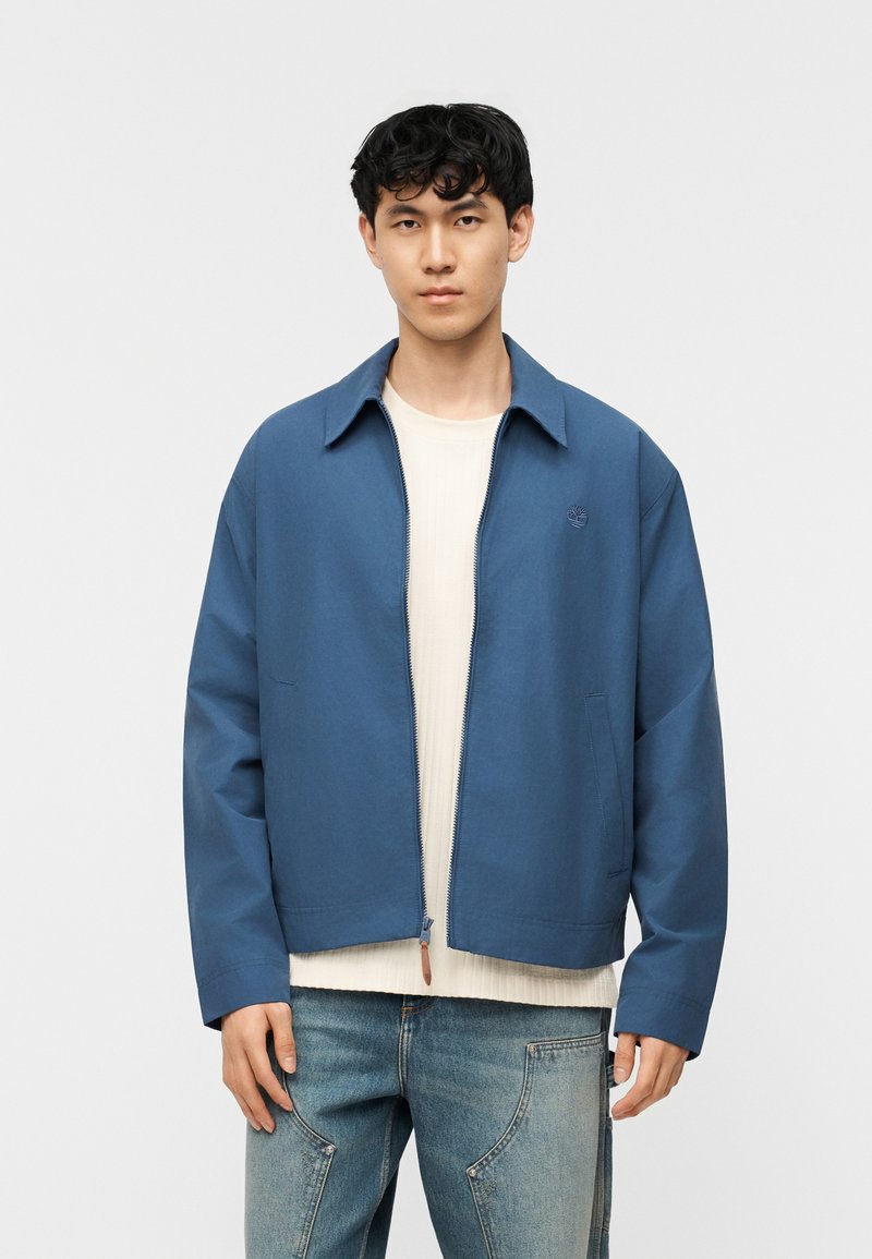 Νέος άνδρας που φοράει μπλε μπουφάν με φερμουάρ πάνω από λευκό ριπτό πουκάμισο και ανοιχτόχρωμα τζιν, στέκεται μπροστά σε ένα απλό ανοιχτό φόντο.