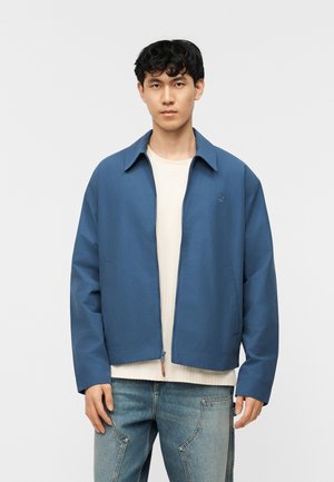Ung mand iført en blå lynlåstrøje over en hvid ribbet skjorte og lyseblå jeans, stående mod en ensfarvet lys baggrund.
