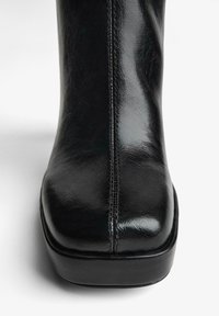 Cizmă neagră de gleznă din piele, cu finisaj lucios, vârf pătrat, cusături vizibile și toc jos și solid. Textură netedă și accente minimale.