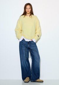 Przewymiarowany, jasnożółty sweter z dzianiny z długimi rękawami, w połączeniu z luźno dopasowanymi niebieskimi dżinsami. Stylizację dopełniają beżowe buty.