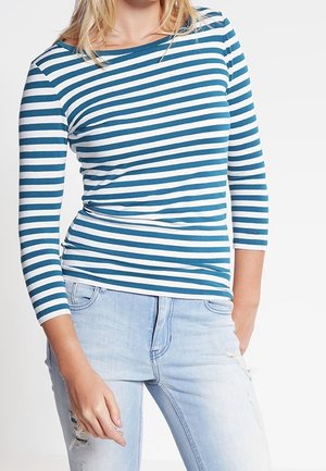 Femme portant un t-shirt à manches longues rayé horizontalement bleu et blanc et un jean déchiré bleu clair, debout avec une main dans la poche.
