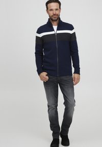 Suéter de punto azul marino con cremallera, con un cuello y puños acanalados, que presenta rayas horizontales en gris y blanco a lo largo del pecho. Combinado con pantalones vaqueros grises.