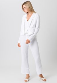 Weißes strukturiertes Strickset mit einem verkürzten Blazer mit Knopfakzenten und passenden Schlaghosen, ergänzt durch weiße Rutschsandalen.
