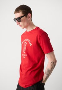 Rød bomulds t-shirt med hvid tekst og logo design, parret med sorte firkantede solbriller. Har korte ærmer og rund halsudskæring.