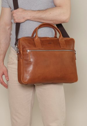 Mann trägt hellgraues Hemd und beige Hose und hält eine hellbraune Lederaktentasche mit Griffen und Schultergurt.