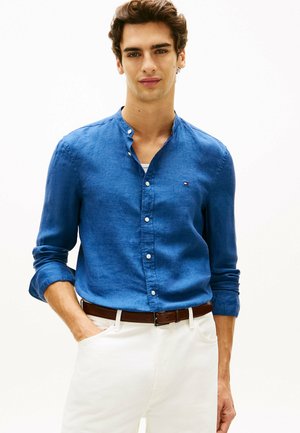 Młody mężczyzna w niebieskiej koszuli na guziki z podwiniętymi rękawami, białych spodniach i brązowym pasku, stoi z jedną ręką w kieszeni.