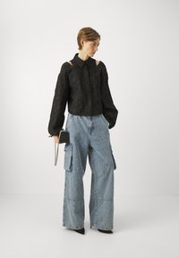 Black cropped blouse with textured floral pattern, paired with oversized blue denim cargo pants featuring pockets. Model holds a small bag.