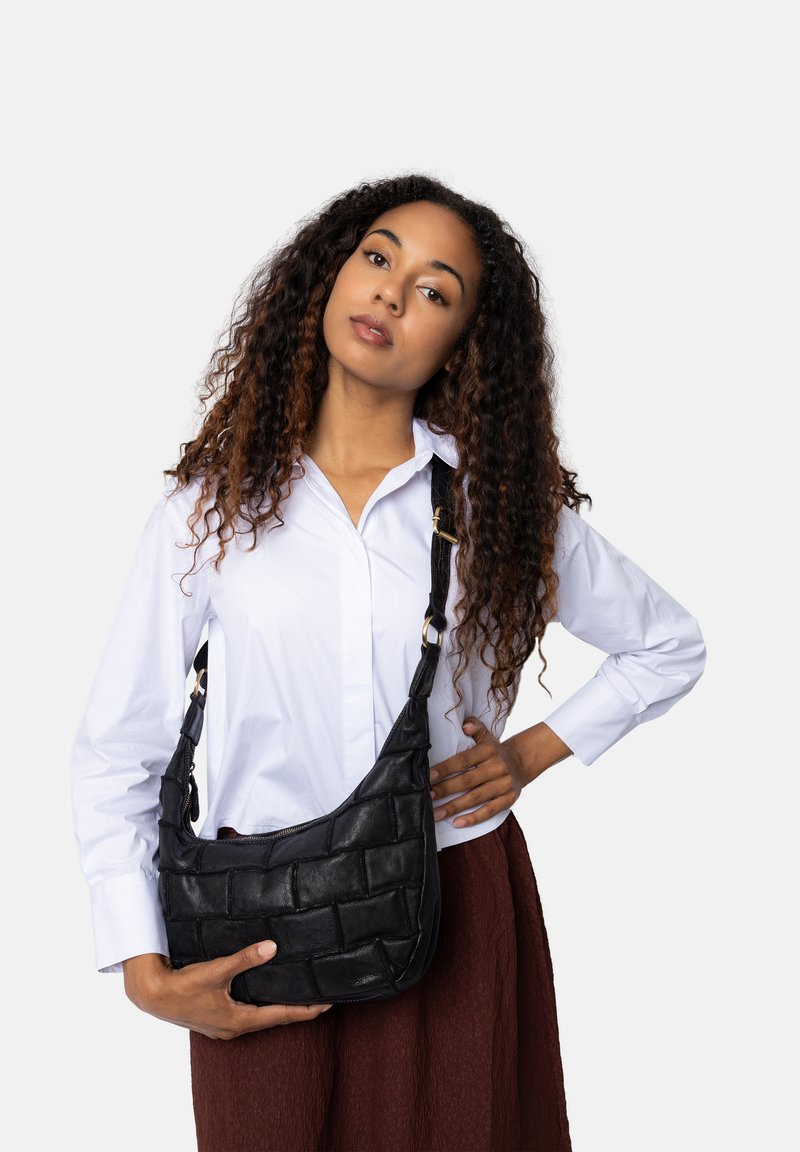 Donna con capelli ricci che indossa una camicia bianca e una gonna marrone tiene una borsa a tracolla nera texture su uno sfondo chiaro uniforme.