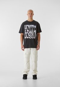 Čierne grafické tričko s nápisom "YOUTH LOST" vytlačeným v skreslených, vlnitých bielych písmenách. Skombinované s krémovými cargo nohavicami a čiernymi teniskami.