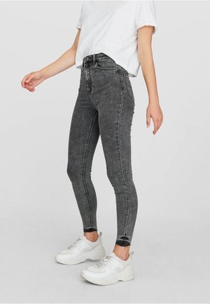 Kobieta w czarnych, wypranych, rurkach z wysokim stanem, białym krótkim T-shircie i białych masywnych sneakersach, stojąca na jednolitym tle.