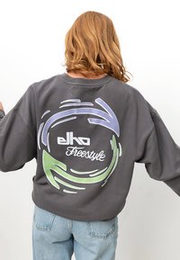 Grauer Sweatshirt mit oversize Passform, ausgestattet mit einem kreisförmigen grafischen Design in Lila und Grün. Der Logo-Text lautet "elko Freestyle." Aus Baumwolle.