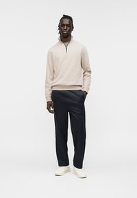 Ανοιχτό μπεζ πουλόβερ με φερμουάρ στο ένα τέταρτο, με ριμπ τελειώματα στα μανίκια, συνδυασμένο με σκούρο ναυτικό μπλε παντελόνι άνετης εφαρμογής και λευκά αθλητικά παπούτσια. Ομαλό ύφασμα με ελάχιστες λεπτομέρειες.