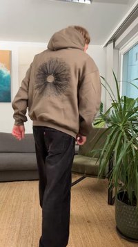 Barna kapucnis pulóver, hátulján kerek fekete grafikával, sötét nadrággal párosítva. Az anyag puha és laza szabásúnak tűnik.