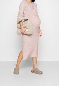 Gravid kvinna i en rosafärgad stickad midiklänning, hållande en beige fluffig väska, iförd grå slip-on skor, stående framför en neutral bakgrund.