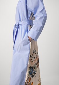 Kobieta ubrana w niebiesko-białą paskowaną sukienkę z paskiem i panelem z kwiatowego materiału, ręka w bocznej kieszeni, na jednolitym tle.