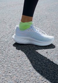 Zapatilla atlética blanca con parte superior de malla texturizada, suela acolchada y acentos reflectantes. Calcetín verde brillante asomando por encima del tobillo.
