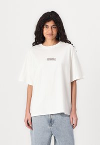 Camiseta blanca de algodón con corte relajado, mangas cortas y escote redondo. Presenta un pequeño logo impreso en la parte frontal en marrón oscuro.