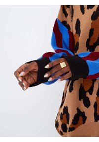 Mains avec des ongles longs blancs portant une bague carrée en or, couvertes par un pull aux motifs léopard bleus, rouges, noirs et bruns.