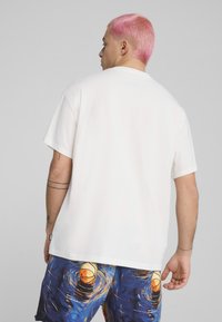 Tricou alb din bumbac cu mâneci scurte, gât rotund și o croială lejeră, asortat cu pantaloni scurți colorați cu modele variate, incluzând forme circulare și ondulate.