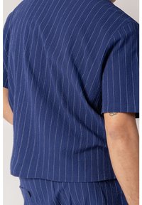 Námořnická modrá košile s krátkým rukávem, vyrobená z texturované látky a s bílými pruhy. Disponuje rovný střihem a pohodlným, uvolněným střihem.